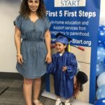 Tall woman in a blue dress holds the hand of a little boy in graduation cap and gown.